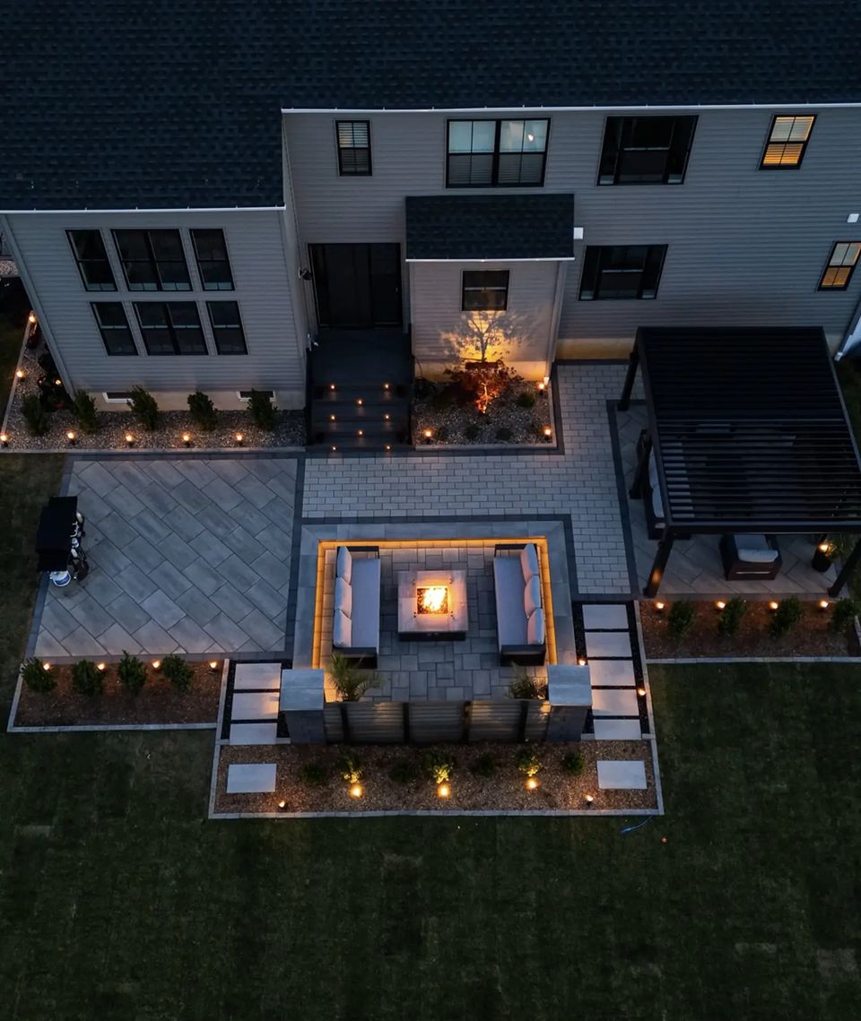 Drone view at dusk of a paver patio with a lit fire table lounge, seat walls and step lights