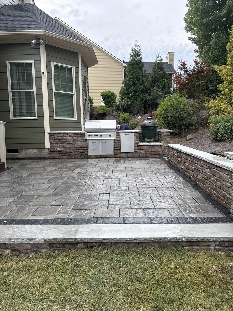 Paver patio with a built in grill and stone seat wall behind a green house