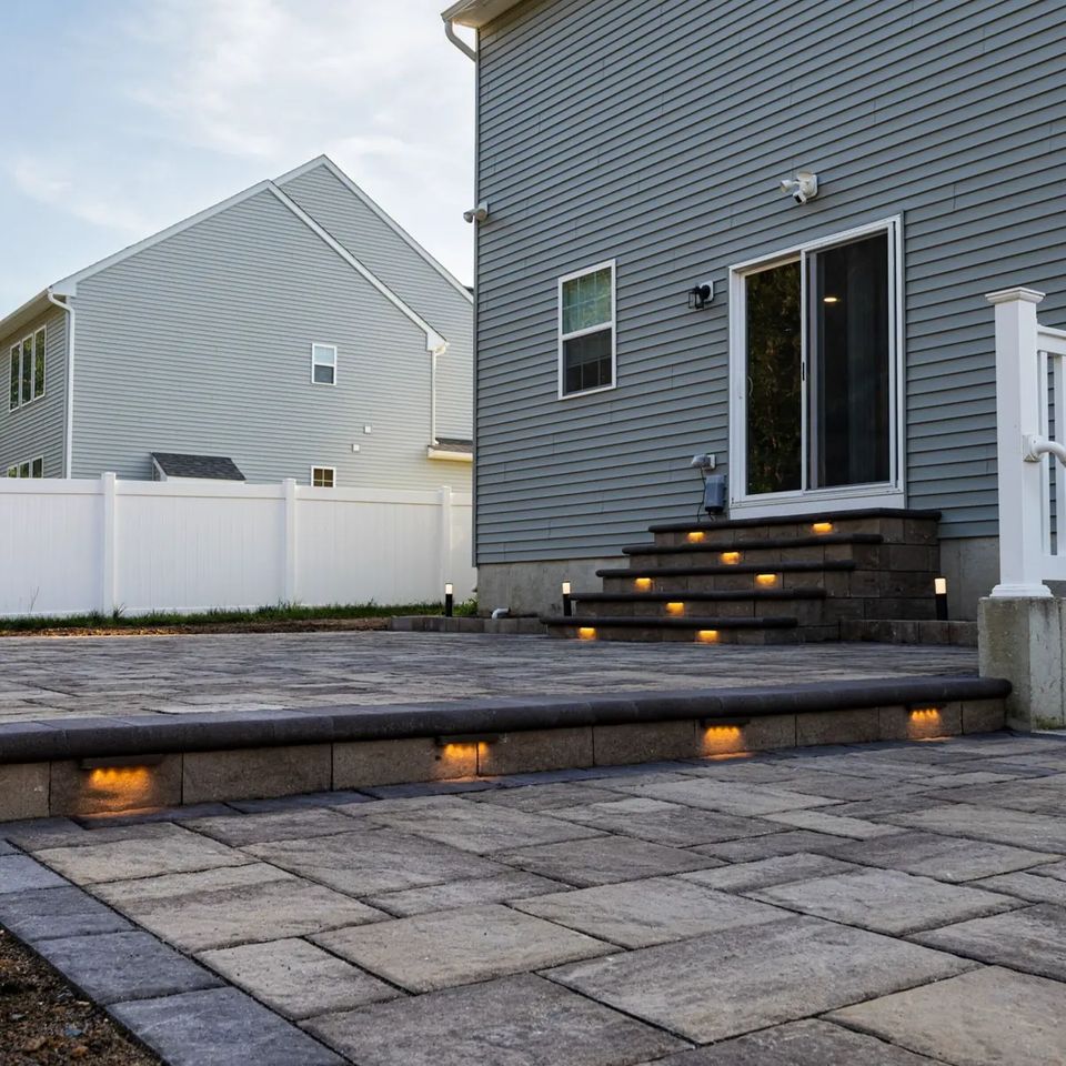 Lit steps down to a paver patio behind a blue gray house